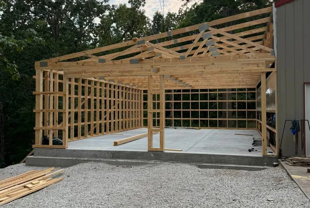 Post-frame extension framing, Alpena AR Post-frame timber framing and roof trusses standing on a slab, joined to the side of an existing cream and red metal building