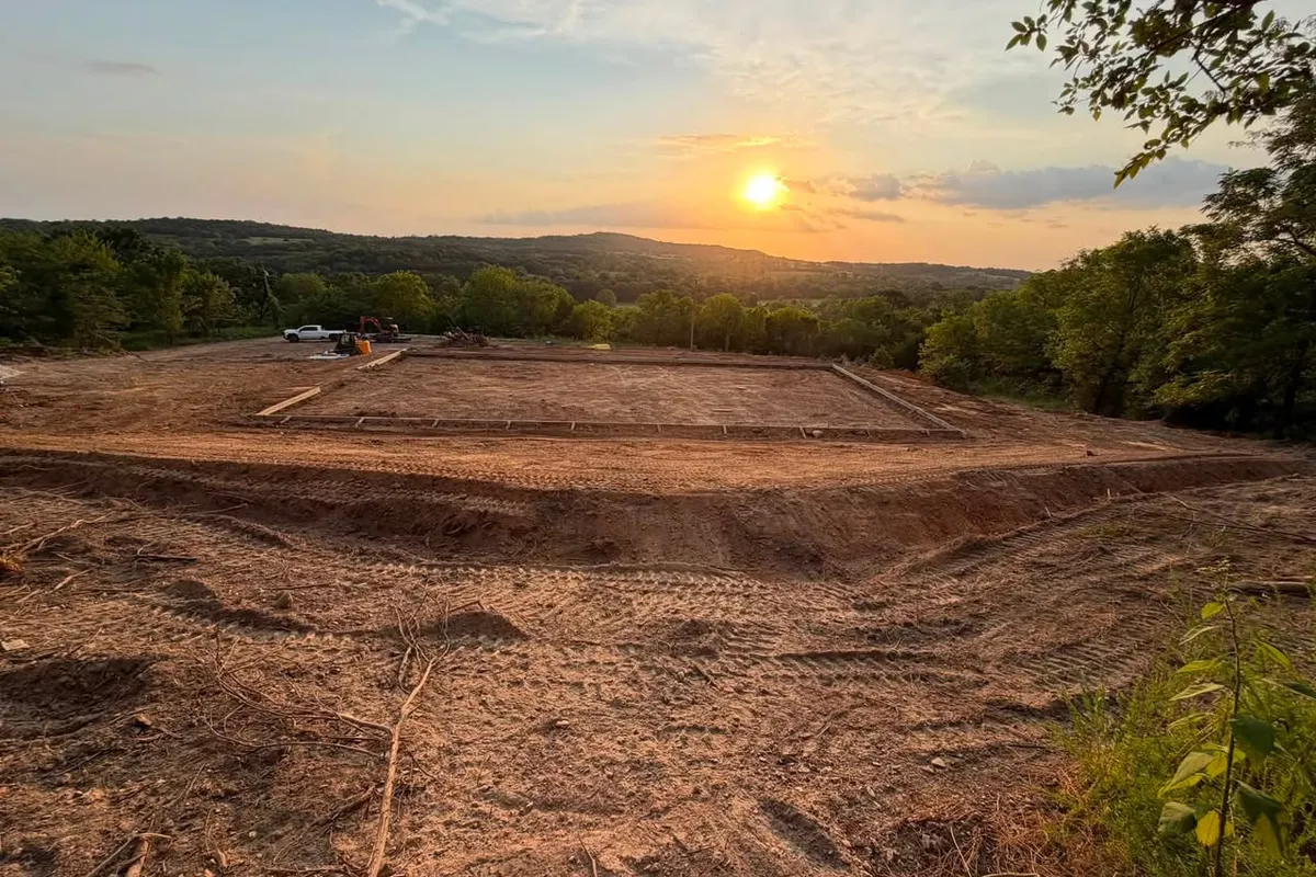 Site preparation, Alpena AR A hilltop building pad cut and levelled at sunset, with footings set out in timber forms and a truck and excavator parked at the far edge