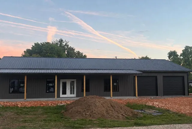 Completed barndominium, Fayetteville AR The finished barndominium at sunset — dark metal walls and roof, white French doors, windows along the covered porch and two closed garage doors at the end