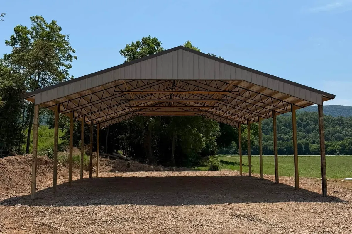 Open-sided equipment shelter, Jasper AR A wide open-sided shelter with timber posts and a brown metal gable roof, standing on gravel with wooded hills behind