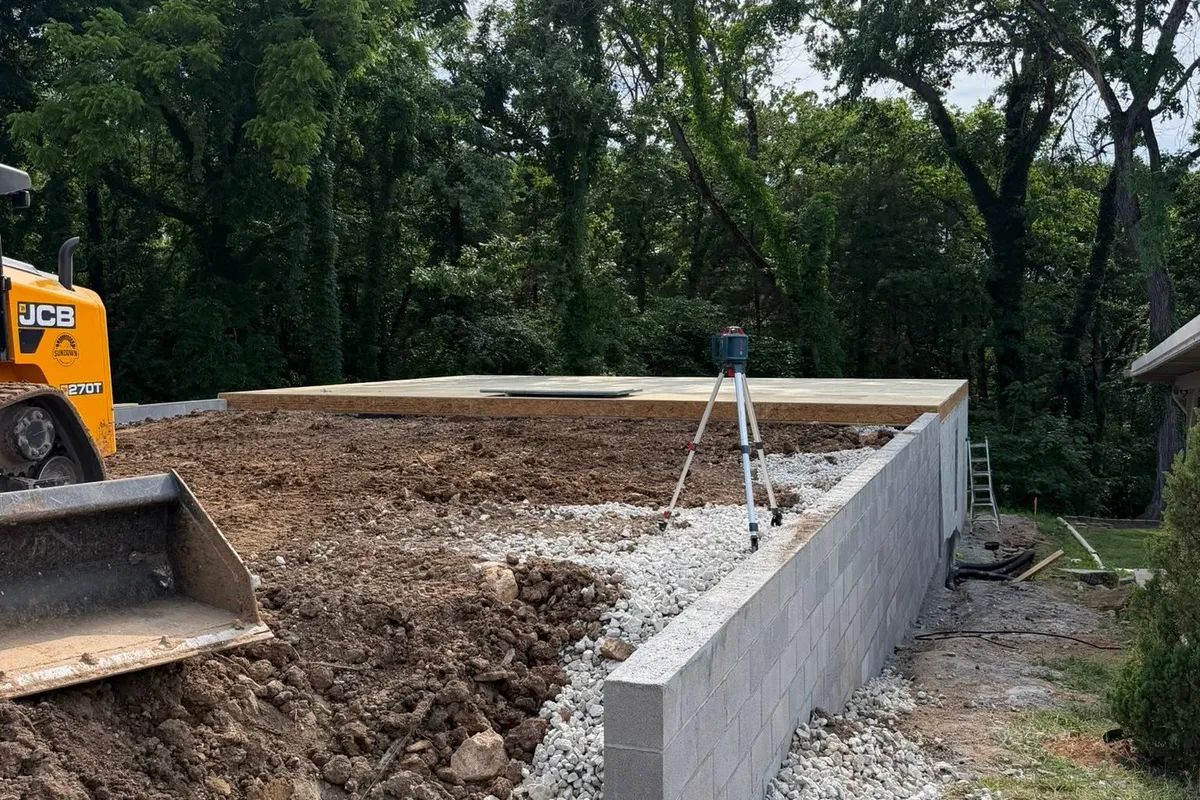 A concrete block stem wall with drainage stone along its base and a subfloor deck behind it, a laser level on a tripod and a telehandler at the edge of the excavation