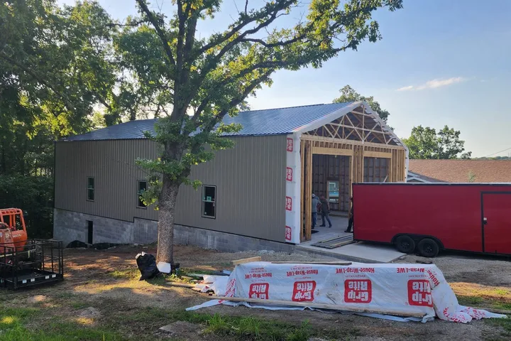 Metal cladding going on, Mountain Home AR The shop clad in olive metal siding with a dark metal roof, the gable end still open and being framed for two door openings, crew working inside