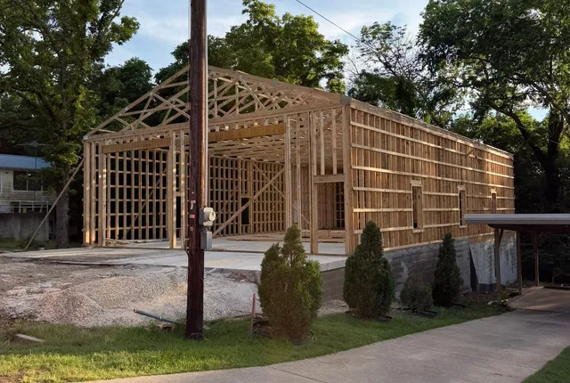 Post-frame timber framing and roof trusses standing on a concrete slab above a block stem wall, beside a residential driveway