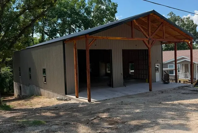 A finished shop in olive metal siding with a dark green roof, stained timber porch posts and gable trusses, and two open bays on a concrete apron