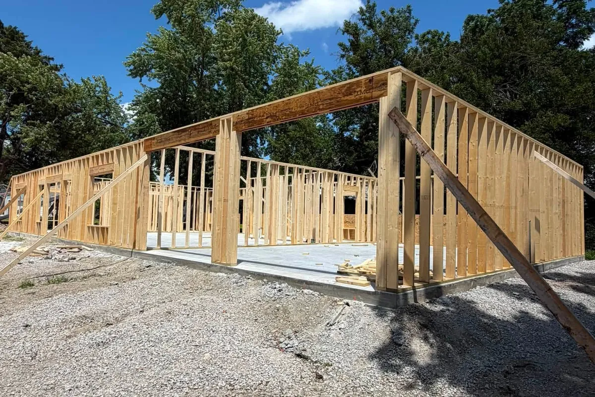 Post-frame timber walls braced and standing on a poured concrete slab over gravel, under a clear blue sky